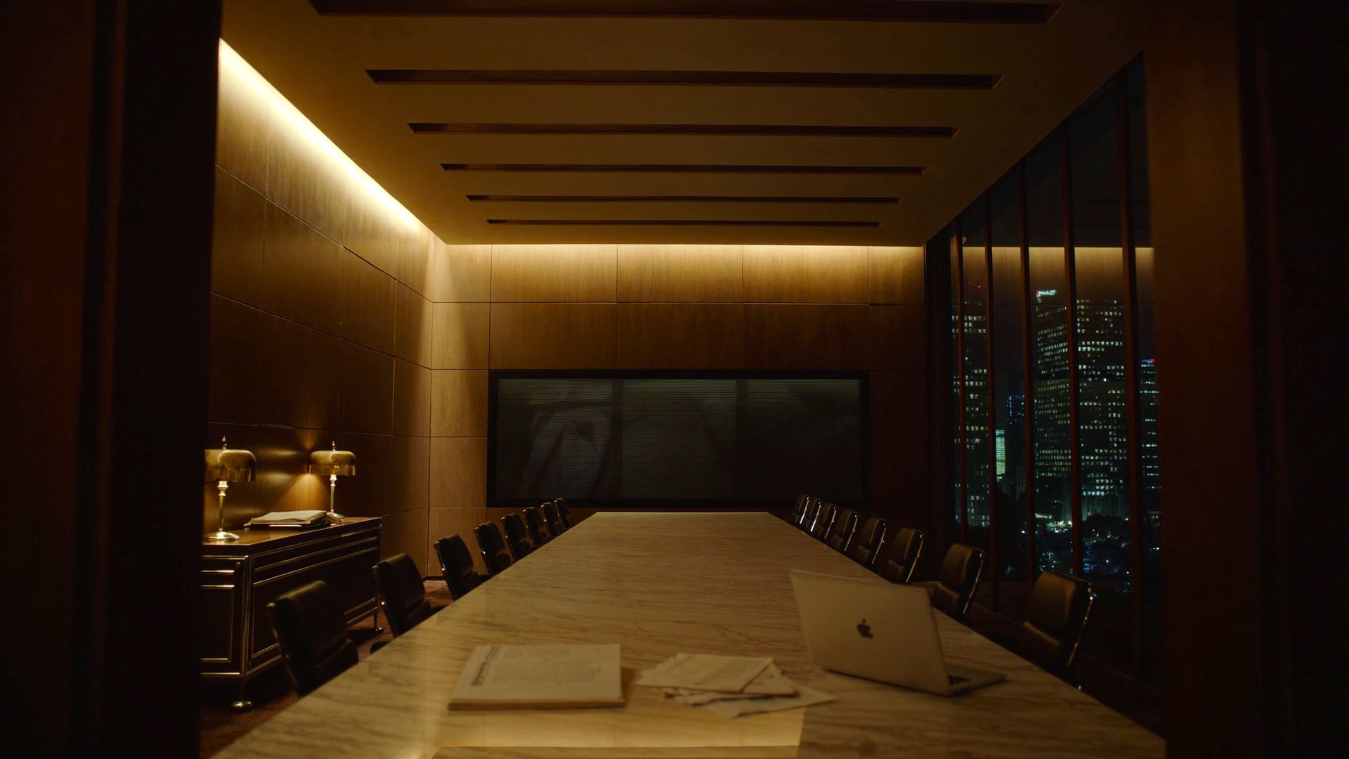 The People in the Loop boardroom at night: a long marble table running toward a dark screen on the wood-panelled back wall, a credenza with two lamps and a stack of books on the left, and the lit city skyline through the window wall on the right.