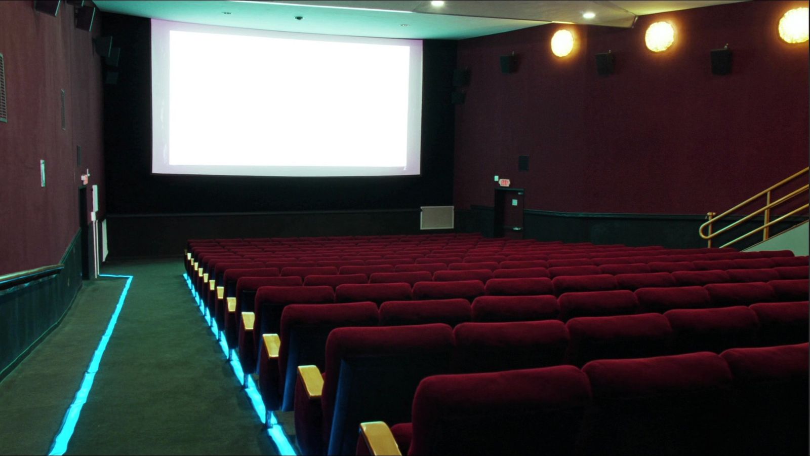 An empty theater with rows of dark red velvet seats, a lit blue aisle strip, and a bright blank screen at the front.